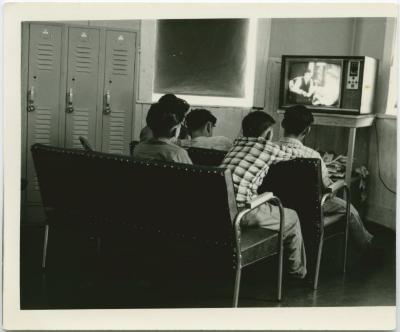 [Male students with back towards the camera watching TV]