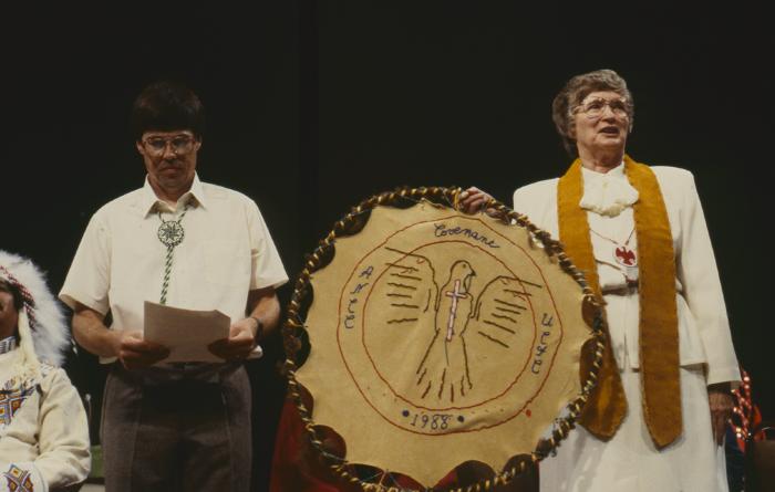 Anne Squire stands facing the council, holding gift presented to the United Church of Canada from the ANCC after the constitution of the ANCC and covenanting service at the 32nd General Council.