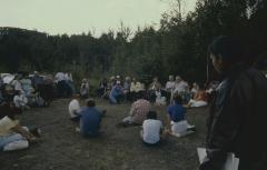 Seated in a circle with Stan McKay in the background are some Native Canadians waiting, contemplating, the apology of the 31st General Council to the Native peoples, in an outdoor setting on the grounds adjacent to the university