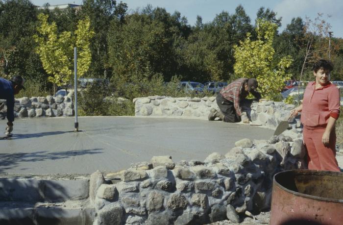 The completed stone wall with Maxine McVey holding the trowel, standing by wall.